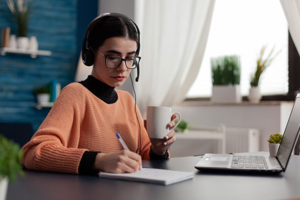 Joven empresaria usando auriculares escuchando los cursos en línea y tomando notas mientras toma café en la sala de su casa