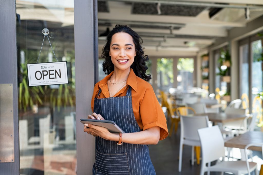 Empresaria mujer dueña de pequeñas empresas o MIPYME en la entrada de su establecimiento de café con una tableta digital.