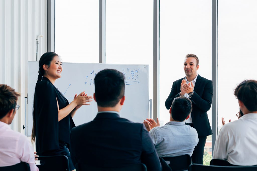 Empresarios levantando la mano en el evento y un pizarron al frente dando una exposición sobre temas de tendencia en negocios.