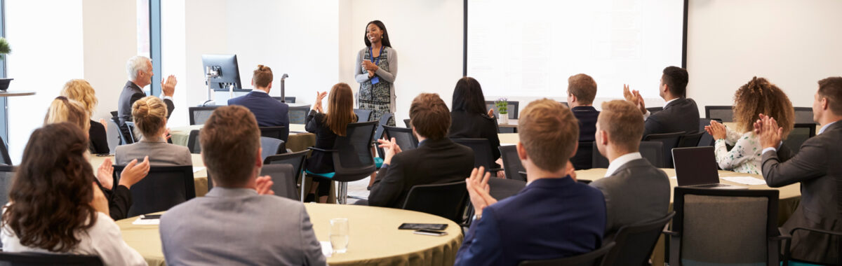 charlas-para-empresarios-1 Expositora comparte ideas clave en charlas para empresarios, con público atento y disposición de mesas redondas tipo conferencia.