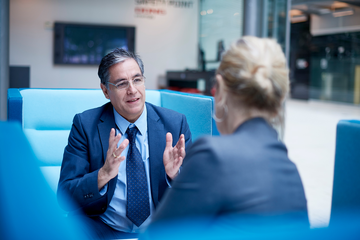 small-business-development-center-1 Hombre de traje hablando con mujer en oficina del Small Business Development Center; fondo con mobiliario moderno y azul.