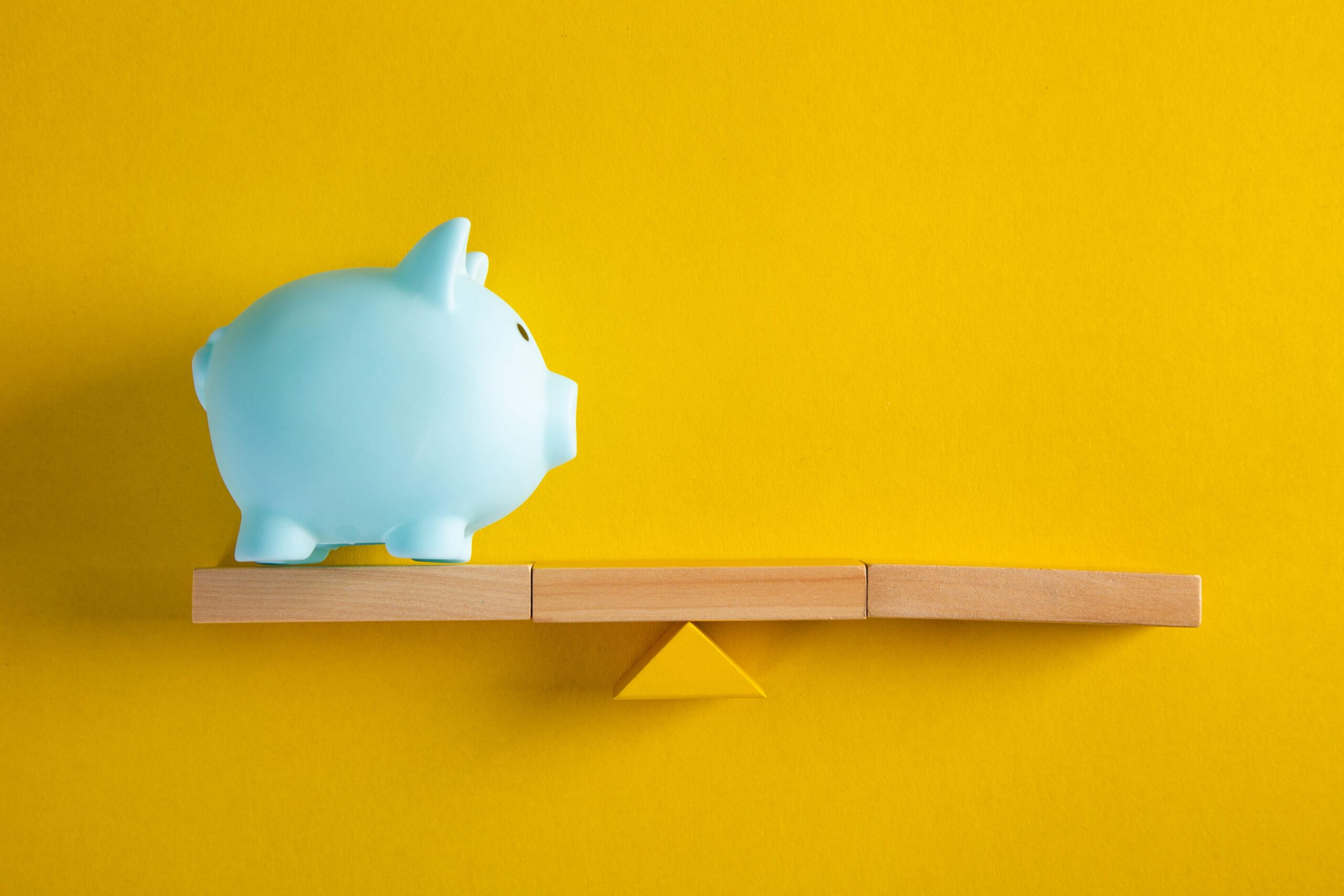 Cómo se interpreta un balance general de los ahorros Una alcancía de cerdito azul sobre una balanza amarilla, ilustrando cómo se interpreta un balance general.